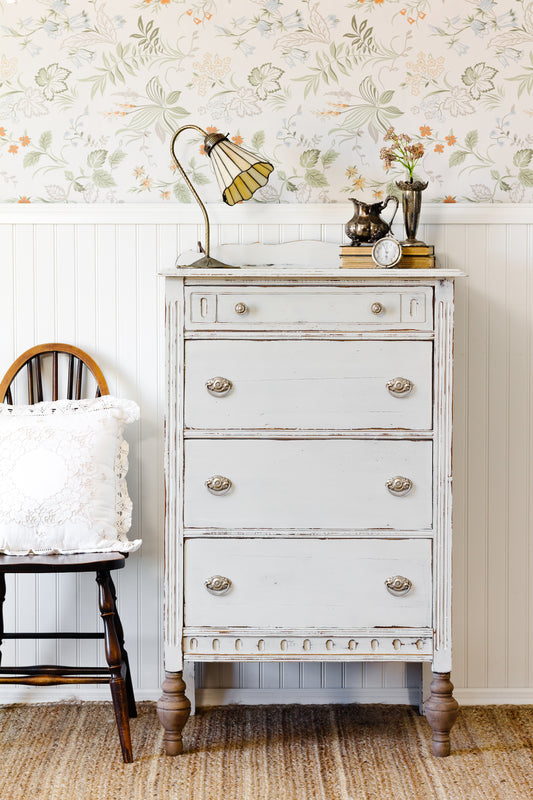 Pale Grey distressed dresser painted in Homestead House Milk Paint with decorative items against a floral wallpapered wall.