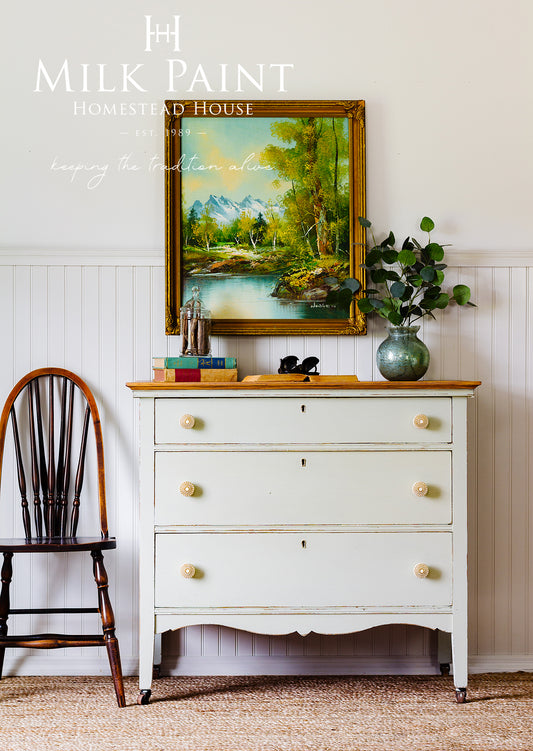 Pot pourri homestead house milk paint chestbof drawers with casters and gold knobs, a wooden chair, and a landscape painting on a white wall.