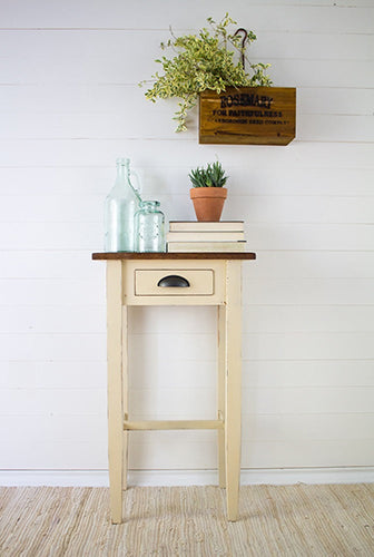 Cream small wooden table with a drawer painted in Buttermilk Cream Homestead House Milk Paint, dressed with books and plants against a white wall.
