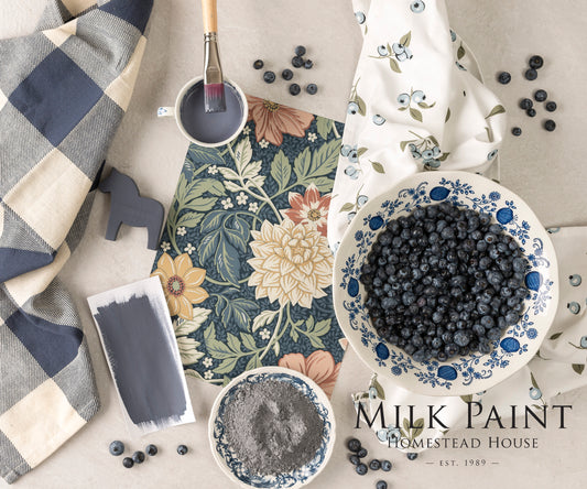 Kurbits Indigo Milk paint inspiration board with painting tools, checked material and floral wallpaper on a surface, featuring Homestead House branding.