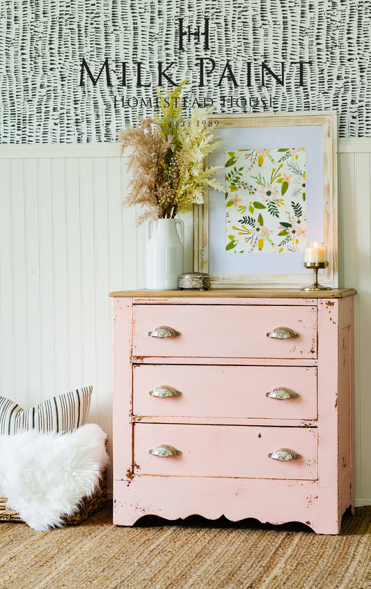 Ronantic feel Homestead House Spring Blossom Milk Paint chest of drawers painted in an old world chippy style with a framed picture and decorative items against a textured wall.