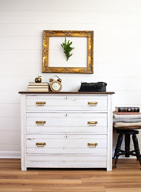 White dresser with gold handles in a room with a plant and books on top