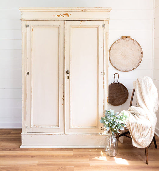 Vintage-style armoire painted in Homestead House Combed Wool Milk Paint in an authentic chippy style in a room with a chair, vase, and decorative items.