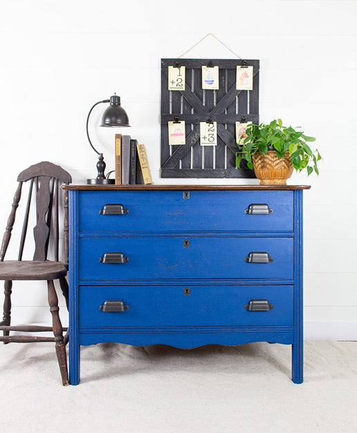 Blue dresser with a wooden top against a white wall, featuring a lamp, books, and a plant.