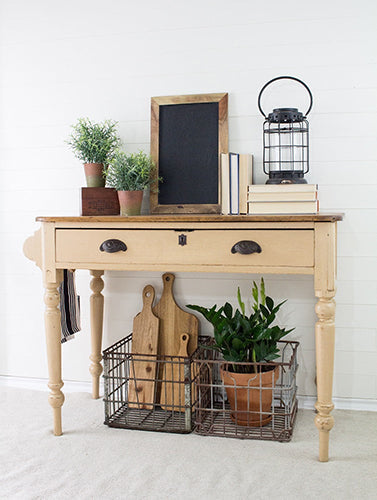 Beige cream wooden console table painted in Hampton homestead House Milk Paint with decorative items including a chalkboard, lantern, and plants against a white wall.